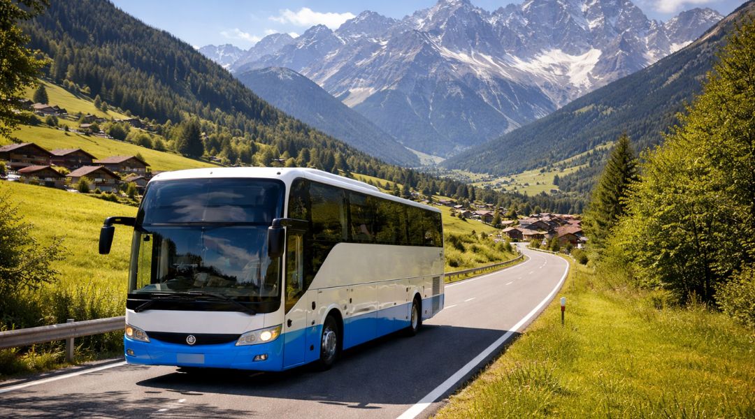 How to Plan a 5 Day Coach Itinerary Through the Austrian Tyrol in 2026. Modern blue and white coach bus driving on a scenic mountain road with lush green valleys, traditional villages, and dramatic snow-capped Alps in the background.