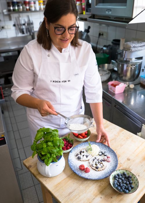 Ein Koch bestäubt einen Dessertteller mit Puderzucker, der mit frischen Beeren dekoriert ist, neben Basilikum und weiteren Früchten in Schalen.