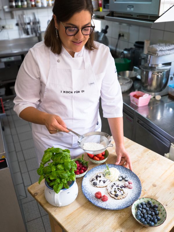 Ein Koch bestäubt einen Dessertteller mit Puderzucker, der mit frischen Beeren dekoriert ist, neben Basilikum und weiteren Früchten in Schalen.