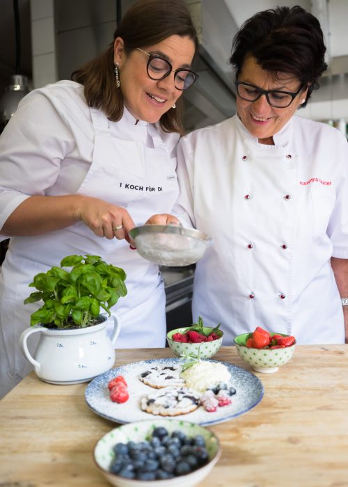 Zwei Köche in weißen Uniformen bereiten ein Dessert mit frischen Beeren und Minze zu, umgeben von einer Auswahl an Früchten und Gebäck auf einem Holztisch.