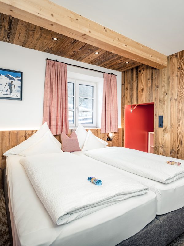 A cozy, rustic bedroom featuring light wood paneled walls, a double bed with white bedding, a teal stool, and pink curtains framing a window.