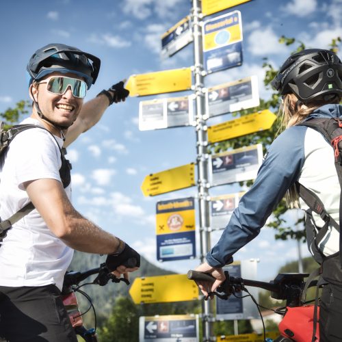 Two mountain bikers wearing helmets and backpacks stand before a trail signpost, with the man pointing toward a direction.